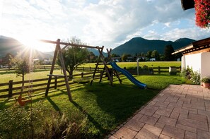 Ein Spielplatz mit Rutsche und Schaukeln, umgeben von einem Holzzaun, einer Rasenfläche und einem Pflasterweg.