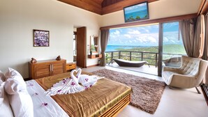 Une chambre spacieuse avec un grand lit, un plafond en bois, une vue sur l’océan, un balcon avec une chaise et un téléviseur fixé au mur.