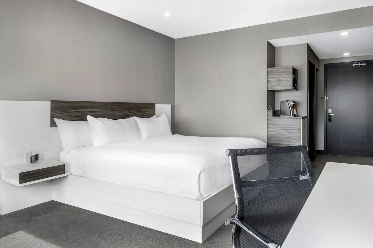 A modern hotel room with a large bed, a bedside table with a lamp, a small seating area, and a door.