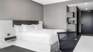 A modern hotel room with a large bed, a bedside table with a lamp, a small seating area, and a door.