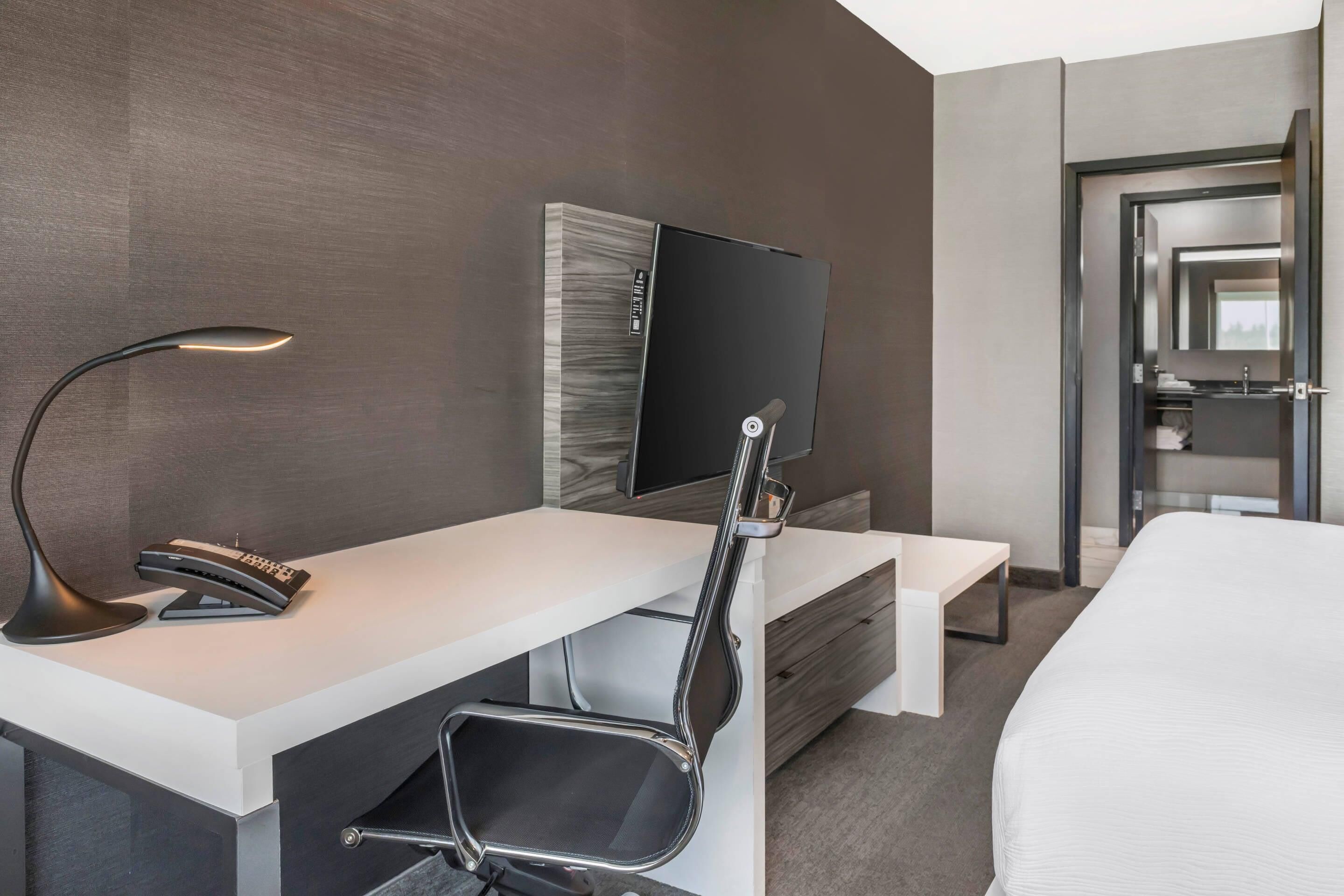 A modern hotel room with a white desk, a black chair, a flat-screen TV mounted on the wall, and a bed with a white mattress.