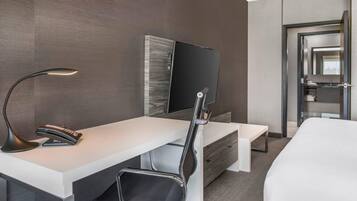 A modern hotel room with a white desk, a black chair, a flat-screen TV mounted on the wall, and a bed with a white mattress.