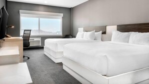 Een moderne hotelkamer met twee bedden, een bureau, een stoel en een groot raam met uitzicht op de stad.