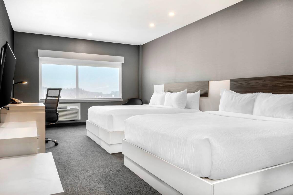 A modern hotel room with two beds, a desk, a chair, and a large window offering a city view.