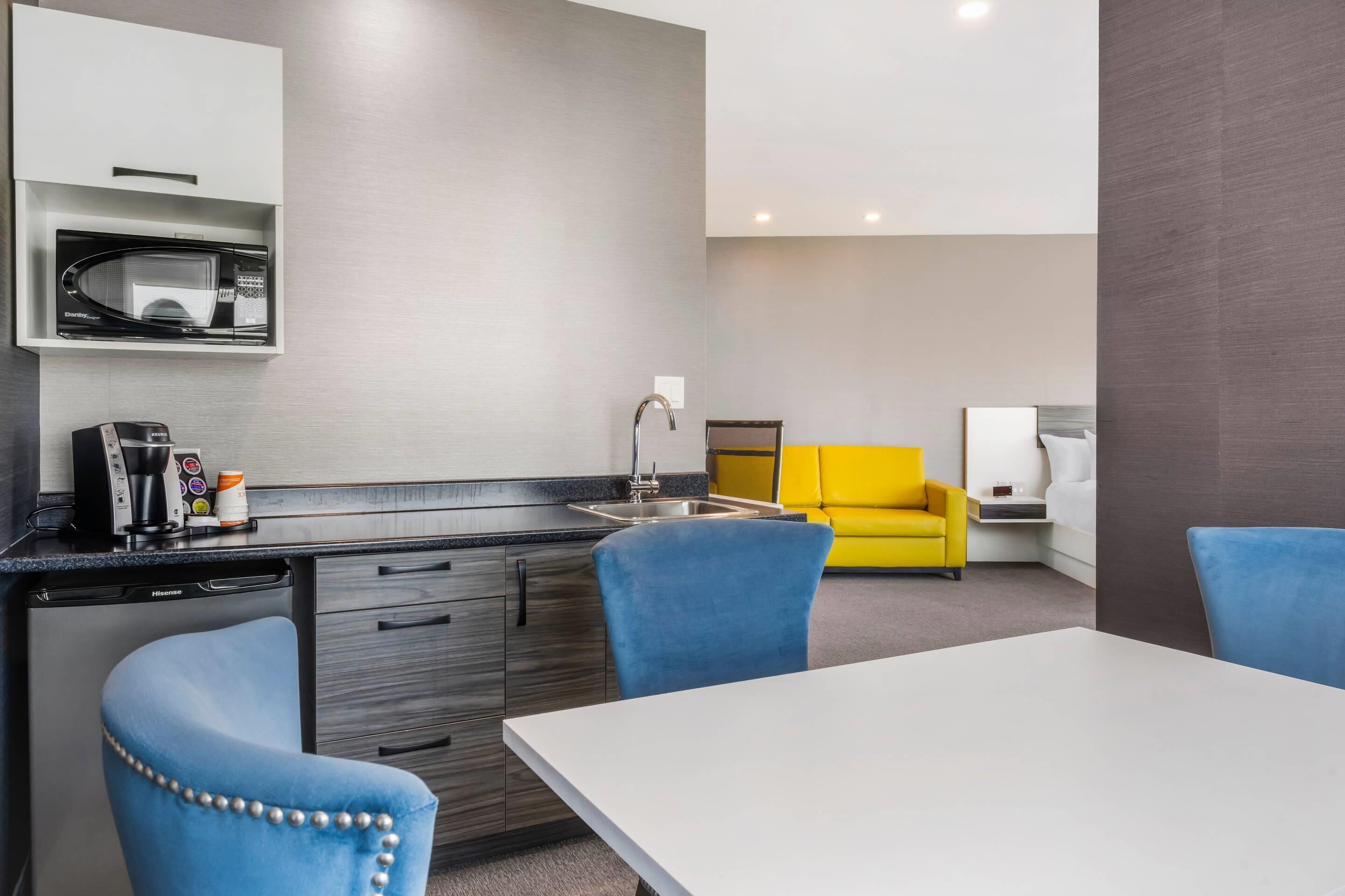 A modern kitchen with a built-in microwave, a coffee maker, and a dishwasher. There is a yellow sofa and a white table with blue chairs.