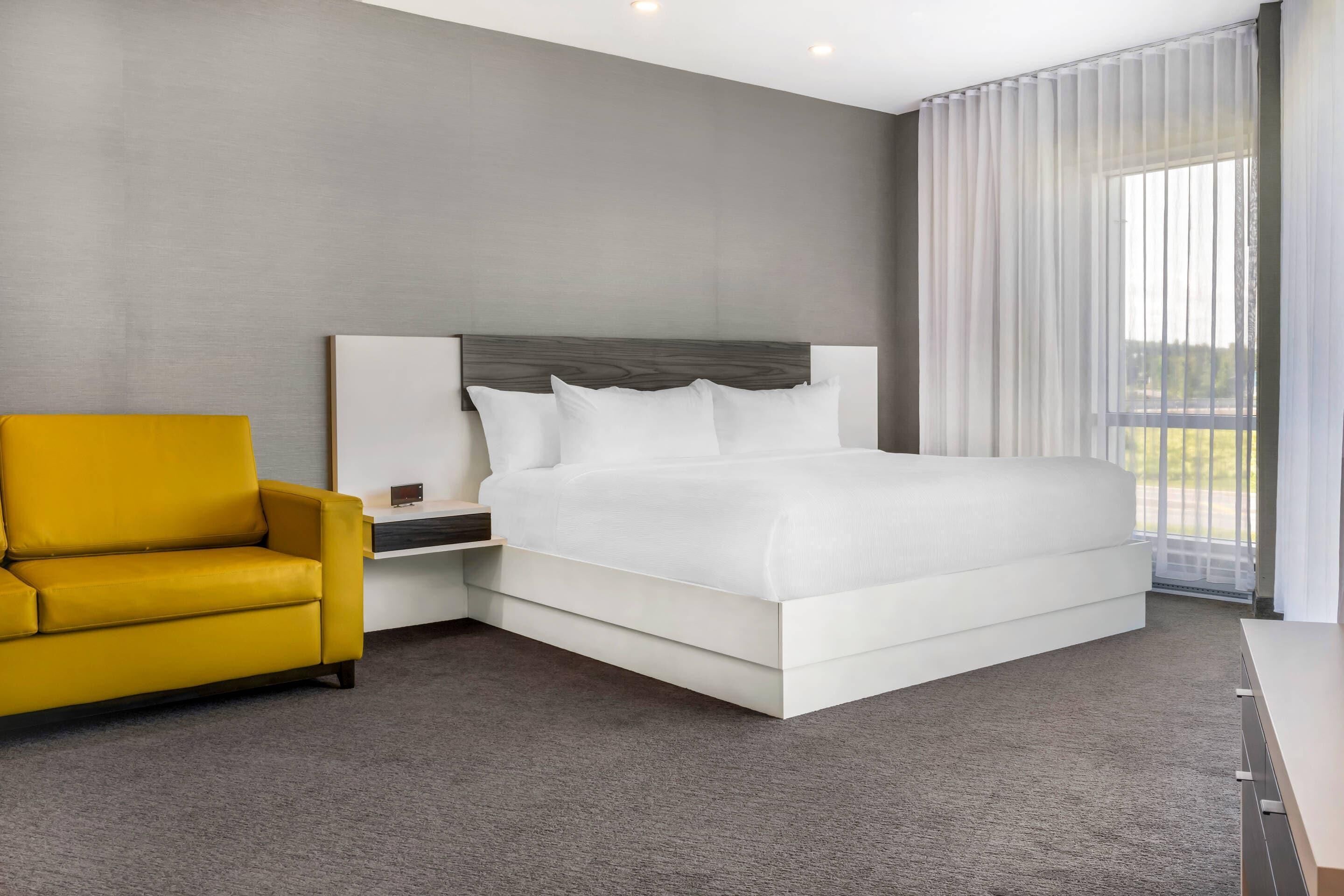 A modern hotel room with a large bed, a yellow armchair, and a view of the outdoors through sheer curtains.