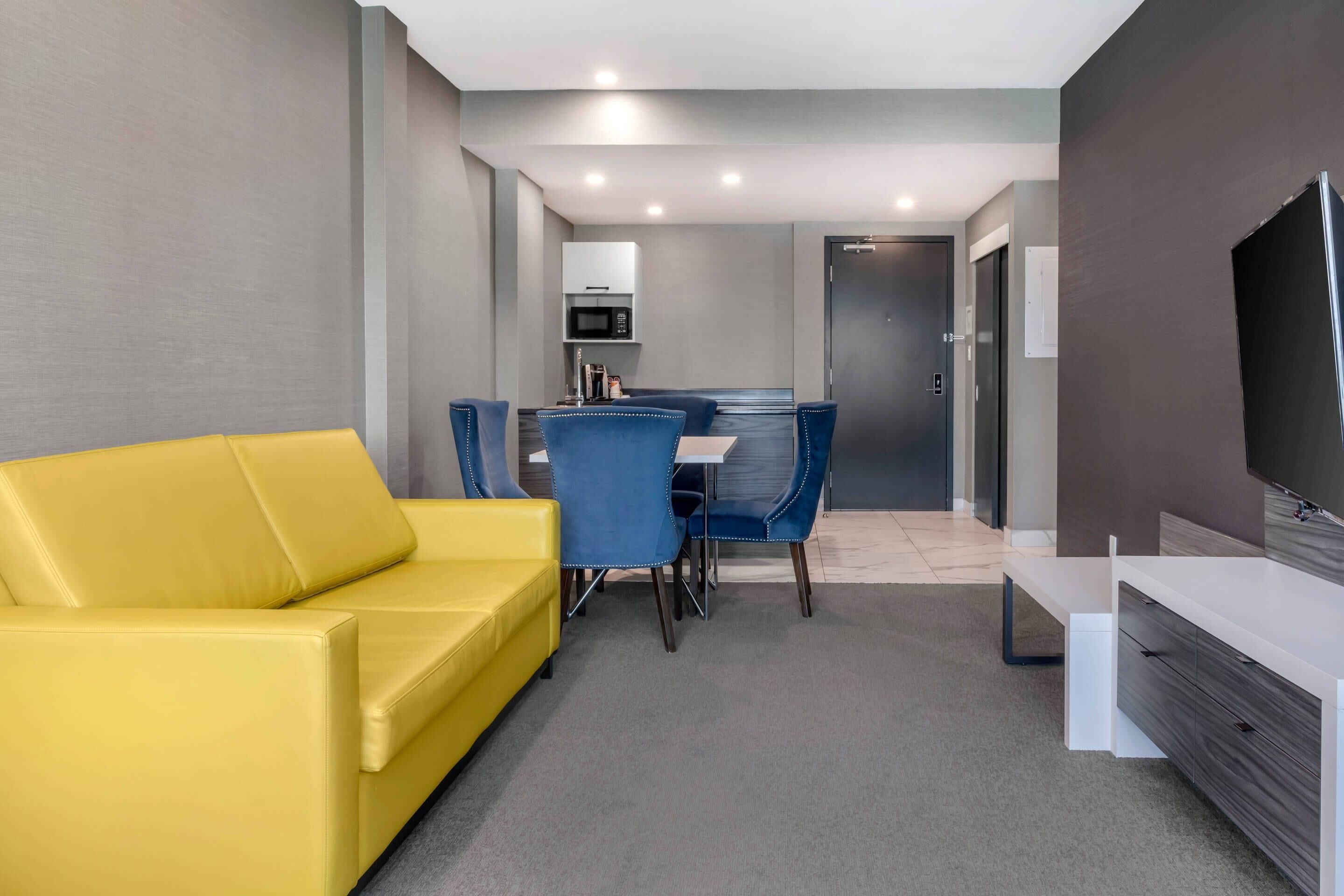 Un salon moderne avec un canapé jaune, une table à manger avec des chaises bleues, un téléviseur à écran plat et un four à micro-ondes en arrière-plan.