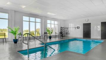 Una piscina interior moderna con agua azul cristalina, rodeada de grandes ventanales, sillas y plantas en macetas.