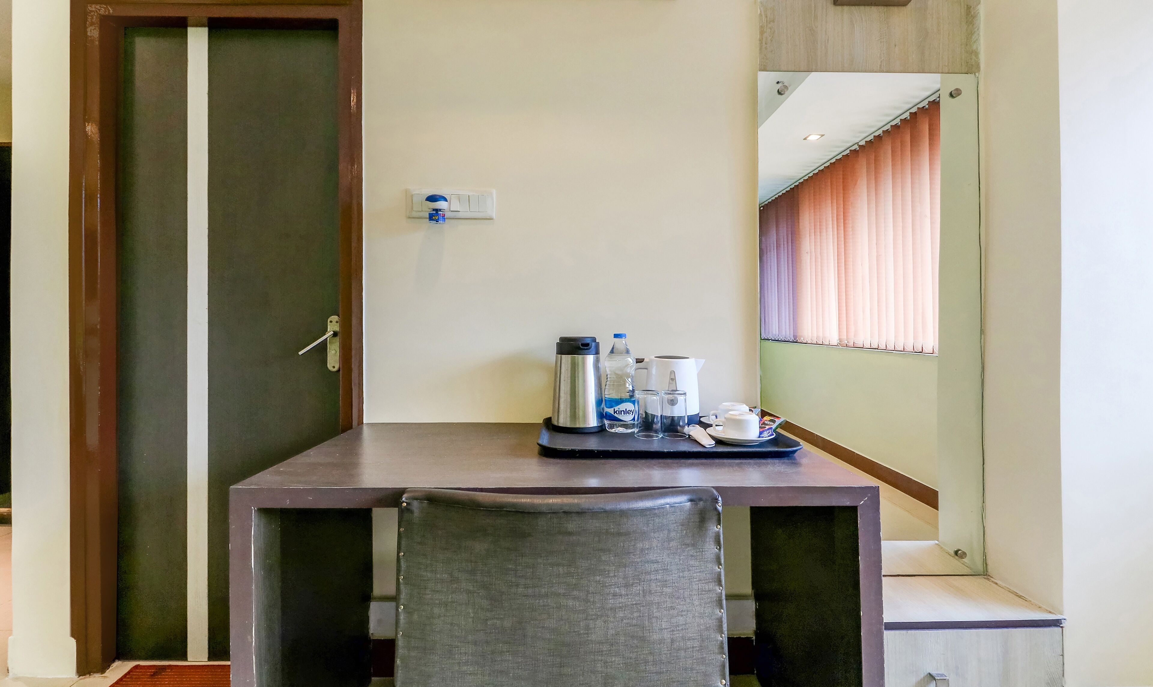 A hotel room with a wooden door, a small table with a water bottle, a thermos, cups, and a tray.