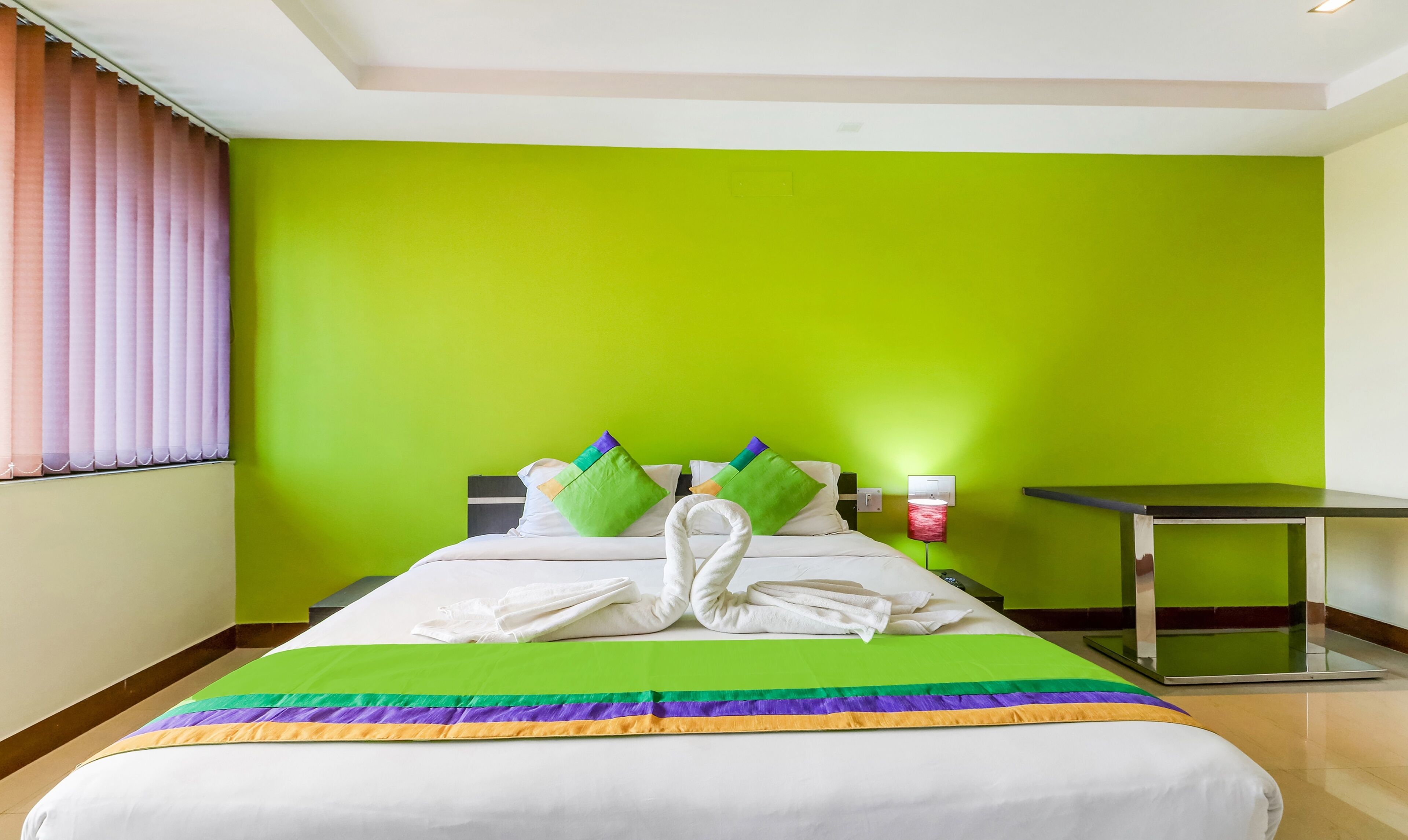 A hotel room with a bed, green walls, a window with curtains, a bedside table, and a lamp.