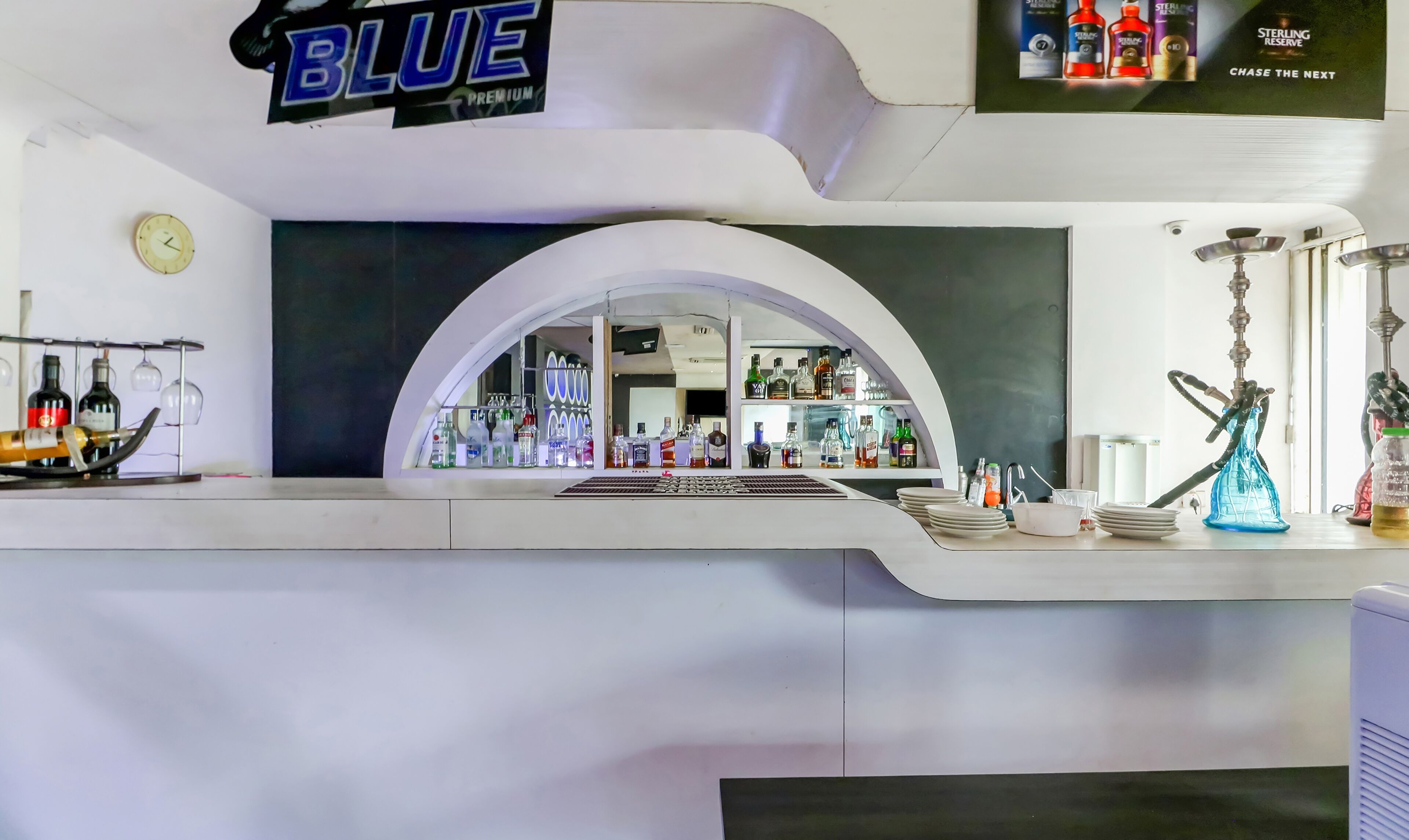 Una zona de bar con diseño moderno, que presenta un llamativo letrero 'BLUE PREMIUM', un reloj en la pared y una variedad de botellas y vasos en estanterías.