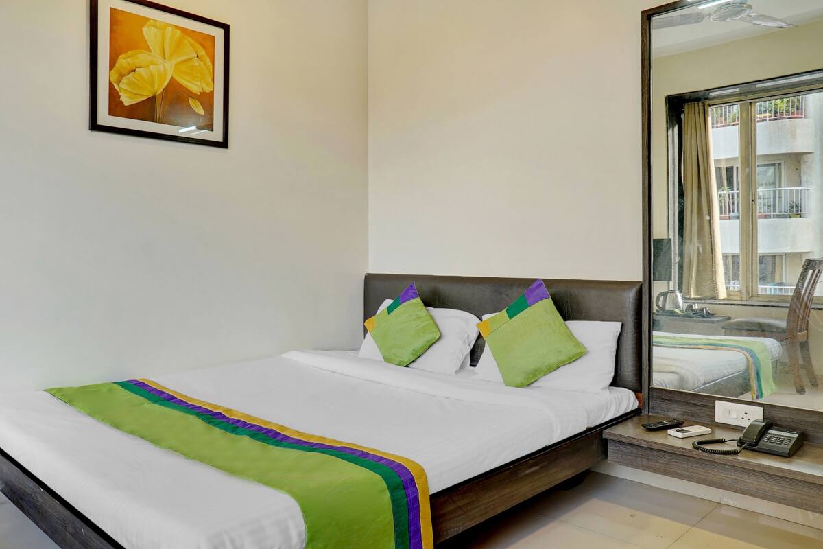 A hotel room with a bed, pillows, a bedside table with a phone and remote, a mirror, and a window with a view of buildings.