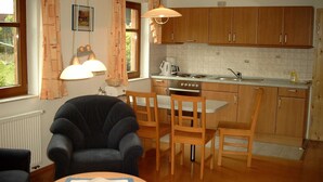 Una cocina compacta con armarios de madera, una mesa de comedor y sillas. Se observa una ventana con cortinas, una lámpara colgante y un radiador.