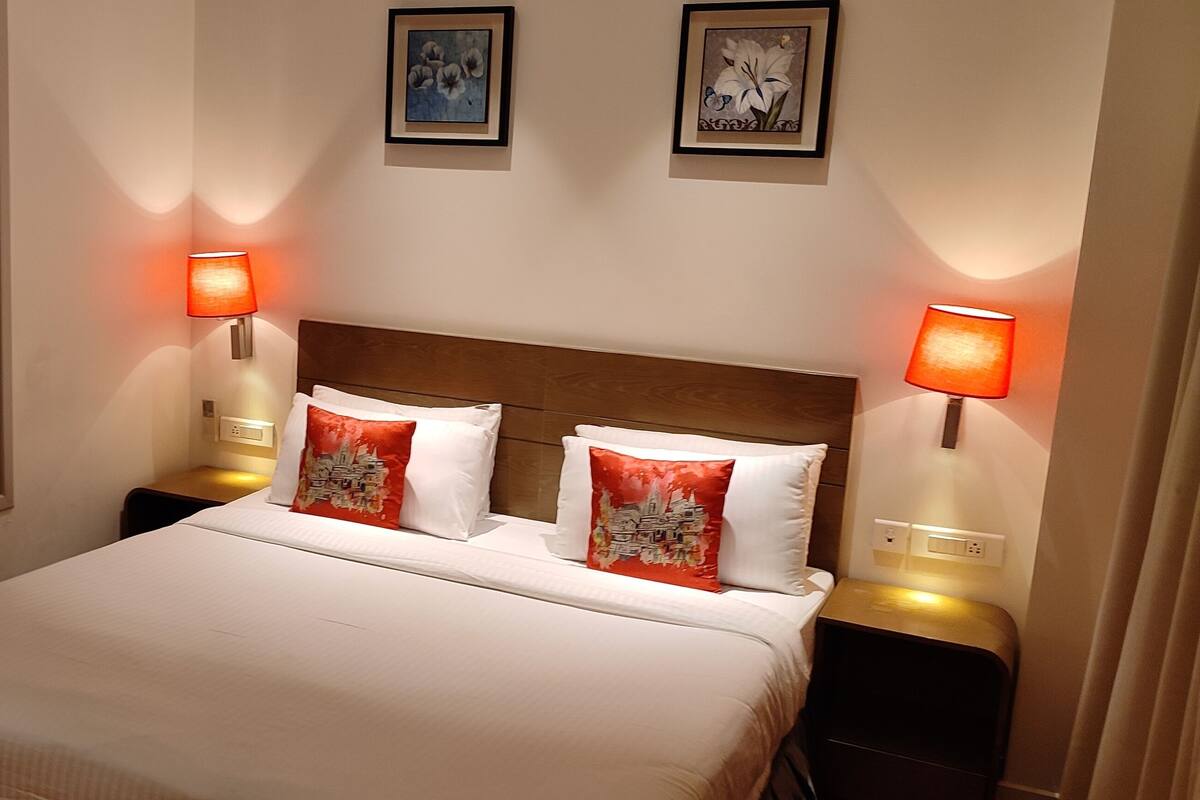 A hotel room with a bed, two bedside lamps, a window, and two framed pictures on the wall.