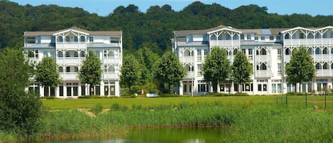 A row of white buildings with multiple stories and balconies, situated next to a body of water and surrounded by greenery.