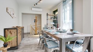 Ein moderner Essbereich mit einem Holztisch, gedeckt für sechs Personen, umgeben von Metallstühlen mit blauen Kissen. Im Hintergrund steht eine hohe Holzskulptur, und eine Lichterkette hängt von der Decke.