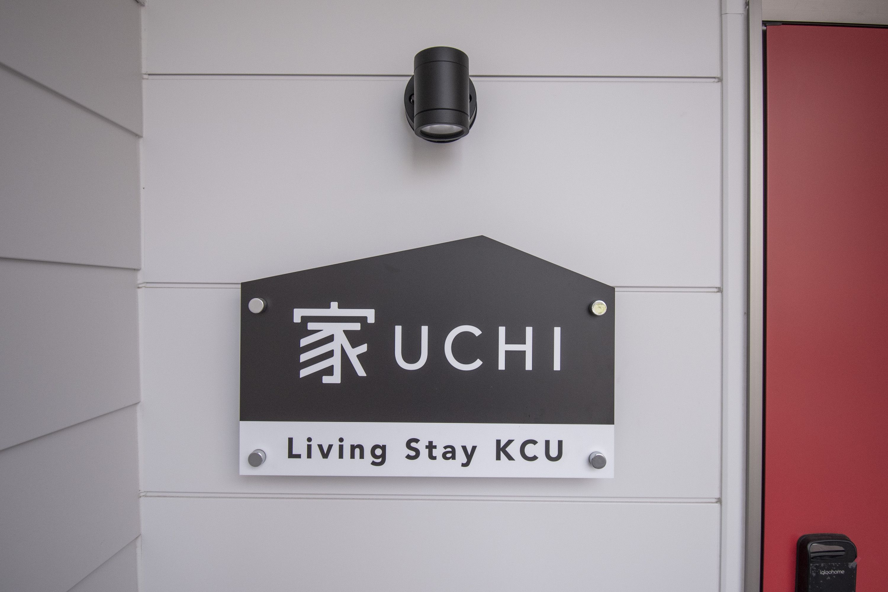 Ein Schild mit dem UCHI Logo und dem Schriftzug "Living Stay KCU", an einer weißen Wand angebracht, mit einer roten Tür in der Nähe.