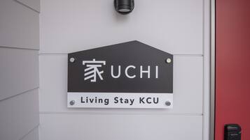 Ein Schild mit dem UCHI Logo und dem Schriftzug "Living Stay KCU", an einer weißen Wand angebracht, mit einer roten Tür in der Nähe.