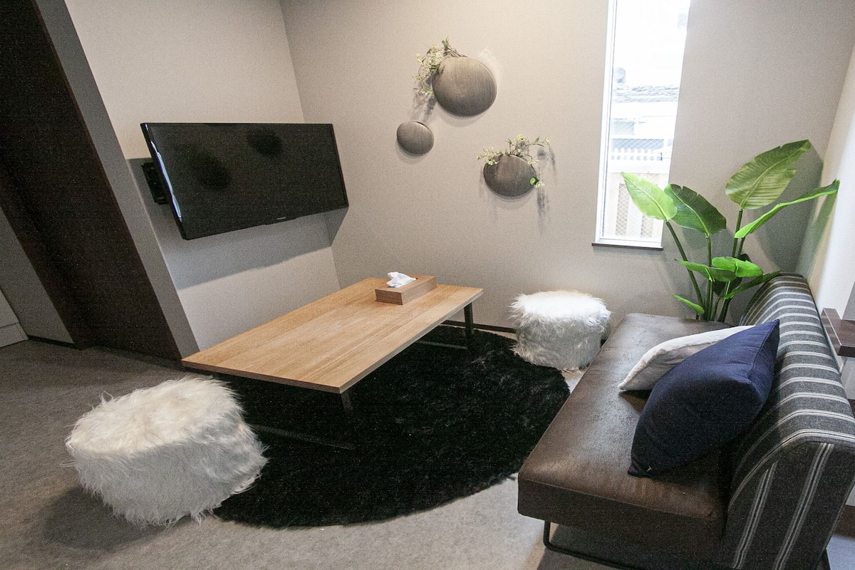 A modern living room with a wooden coffee table, a sofa with cushions, a black furry rug, a TV mounted on the wall, and a potted plant.