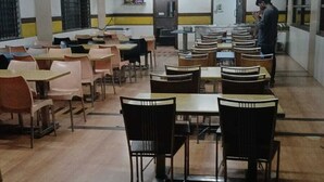 Uma área de refeições com mesas, cadeiras, um ventilador de teto e uma pessoa perto da janela.