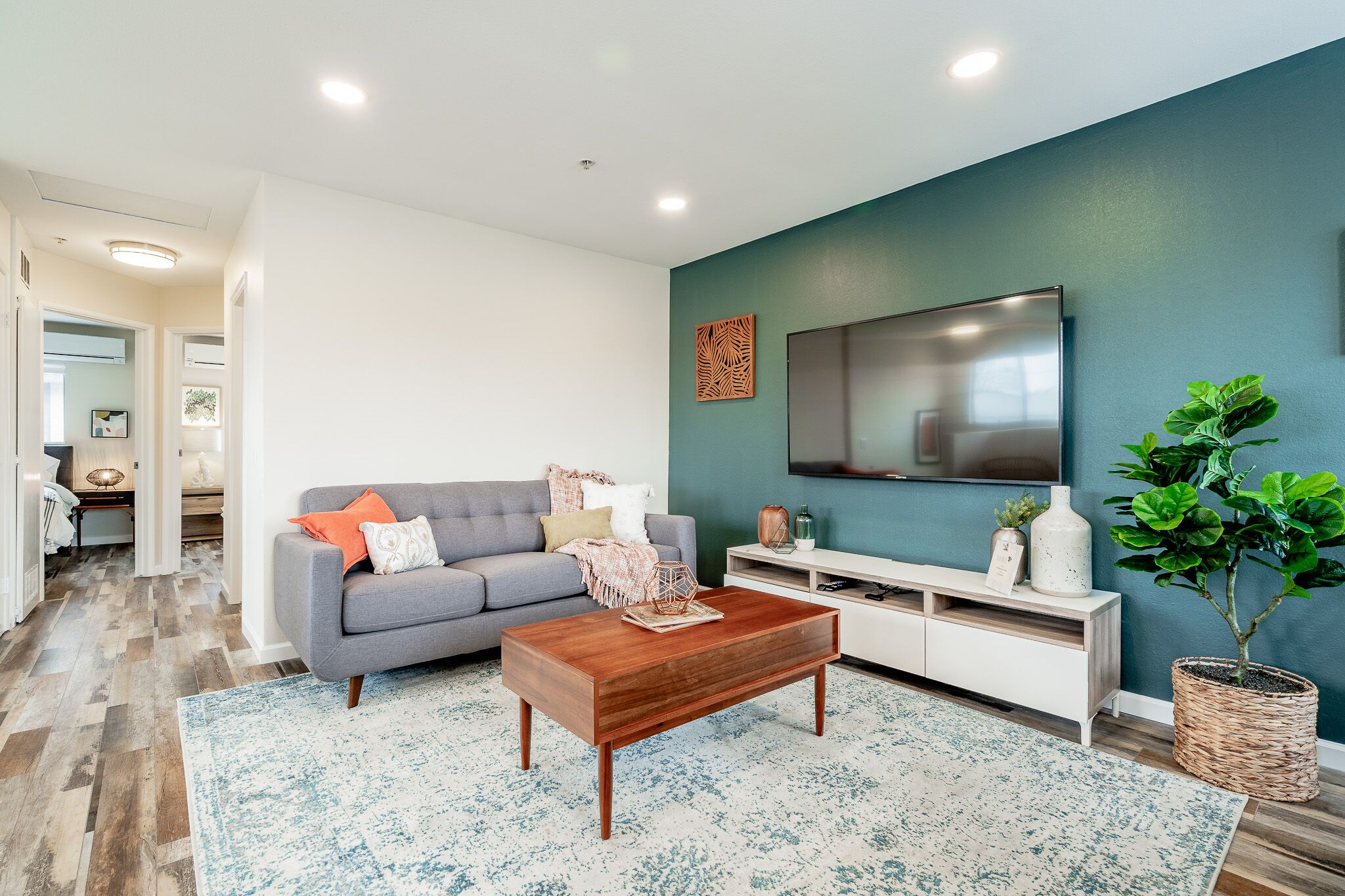 A modern living room with a grey sofa, wooden coffee table, and a large flat-screen TV mounted on a teal wall.
