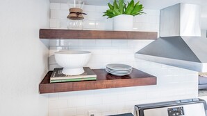 Una cocina con salpicadero de azulejos blancos, estantes de madera, un cuenco blanco, una planta en maceta y una campana extractora moderna.