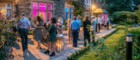 Un bâtiment en pierre doté de nombreuses fenêtres et un espace jardin où se trouvent des personnes rassemblées.