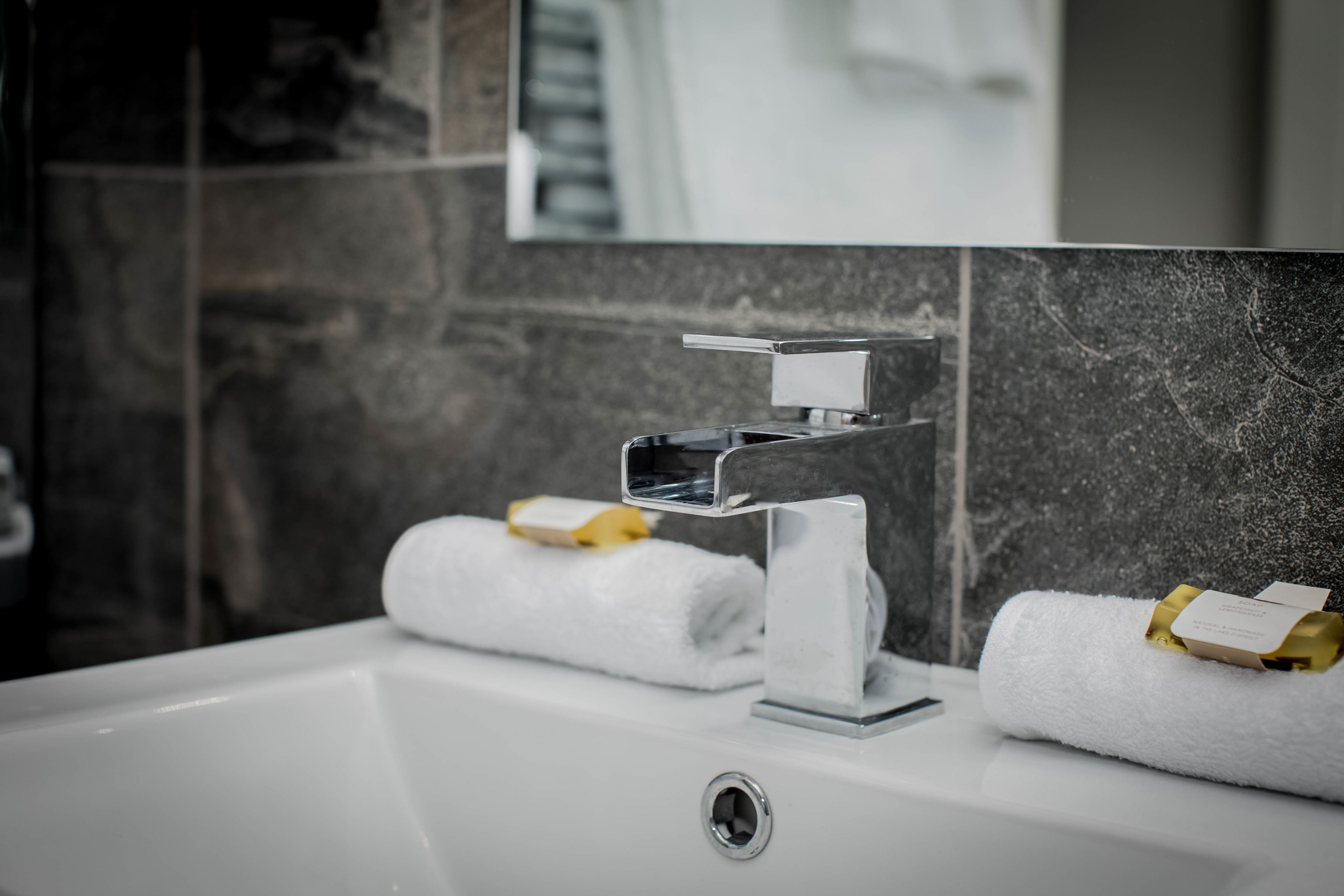 Un baño moderno con un lavamanos blanco, grifo cromado y un espejo encima. Hay dos toallas blancas con detalles dorados sobre el lavamanos.