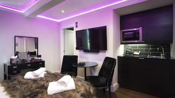 Una habitación de hotel moderna con televisor de pantalla plana, una mesita redonda pequeña, dos sillas negras, un manto de piel, un tocador con espejo y una kitchenette con microondas.