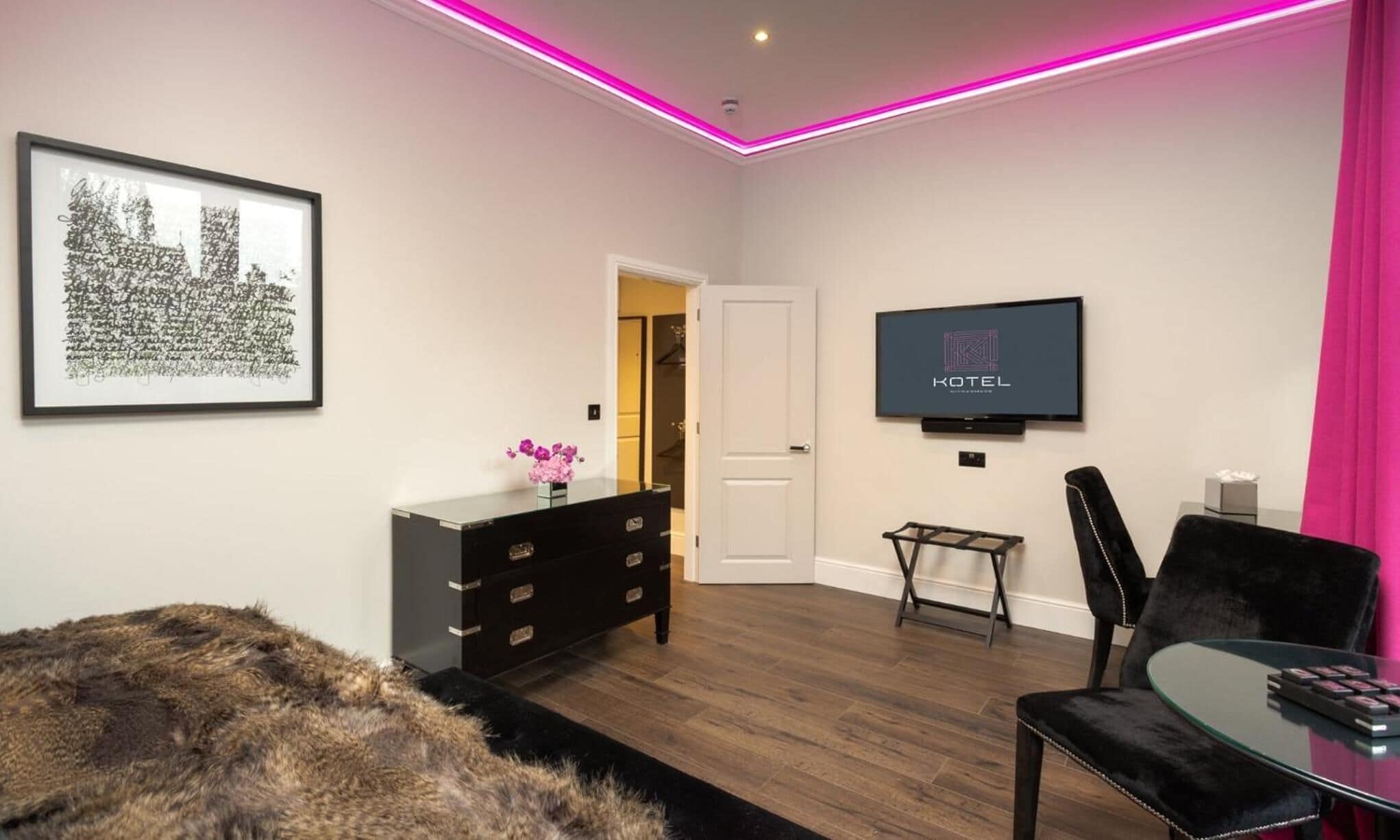 Una habitación de hotel moderna con una televisión de pantalla plana montada en la pared, un sillón de cuero negro, una mesa de centro de vidrio, un manto de piel en el suelo y una obra de arte enmarcada en la pared.