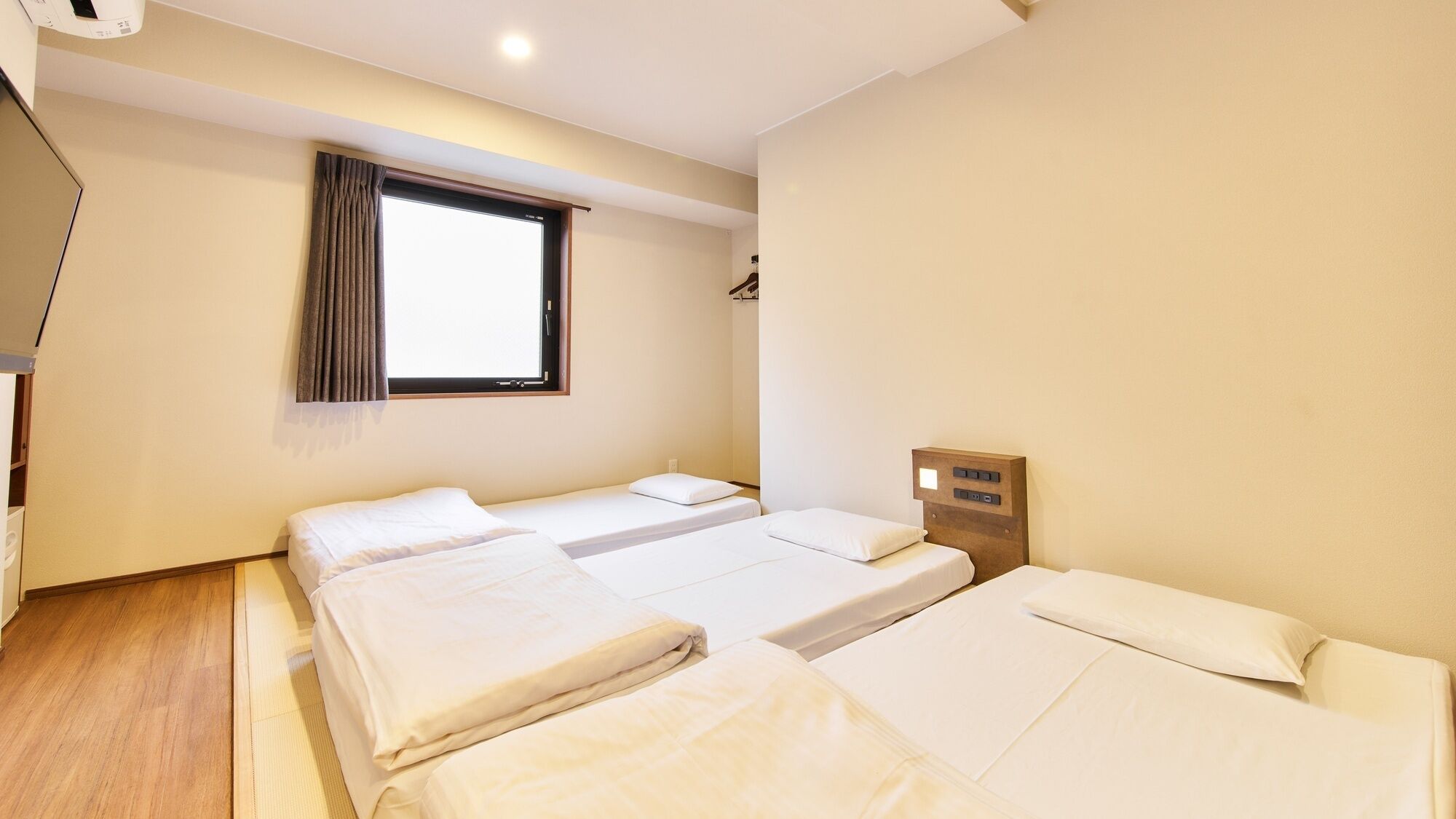 A hotel room with three beds, a window with curtains, a wooden headboard with controls, and a wooden floor.