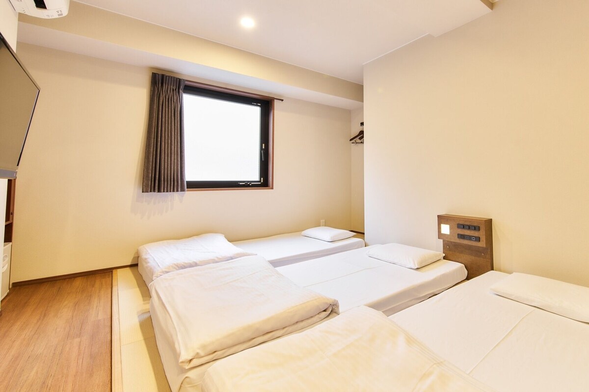 A hotel room with three beds, a window with curtains, a wooden headboard with controls, and a wooden floor.
