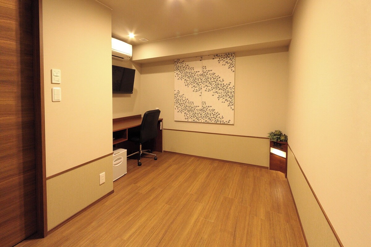 A room with a wooden floor, a desk with a chair, a wall-mounted TV, and a decorative wall piece.