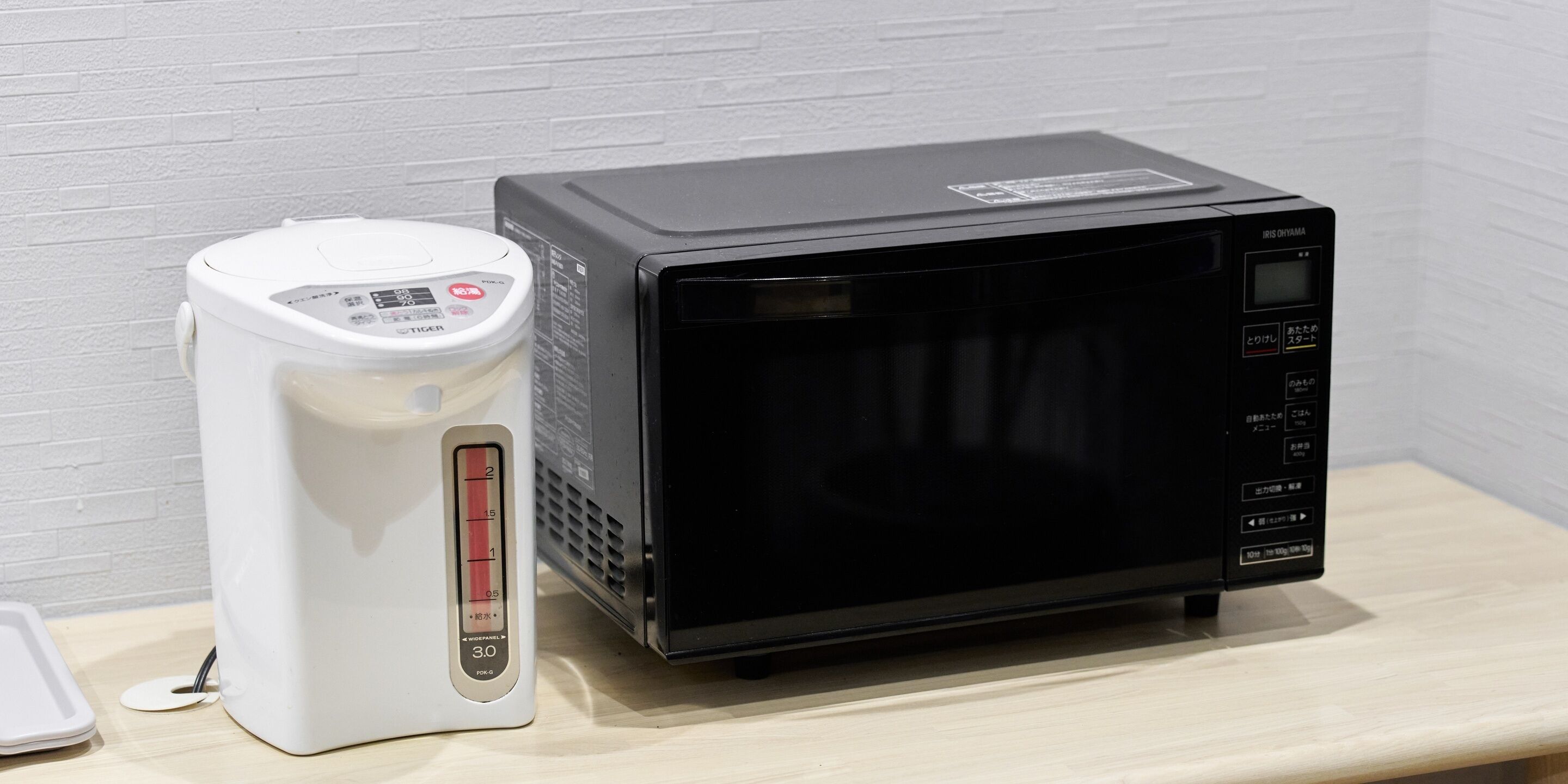 A microwave and a rice cooker on a wooden surface against a white brick wall.