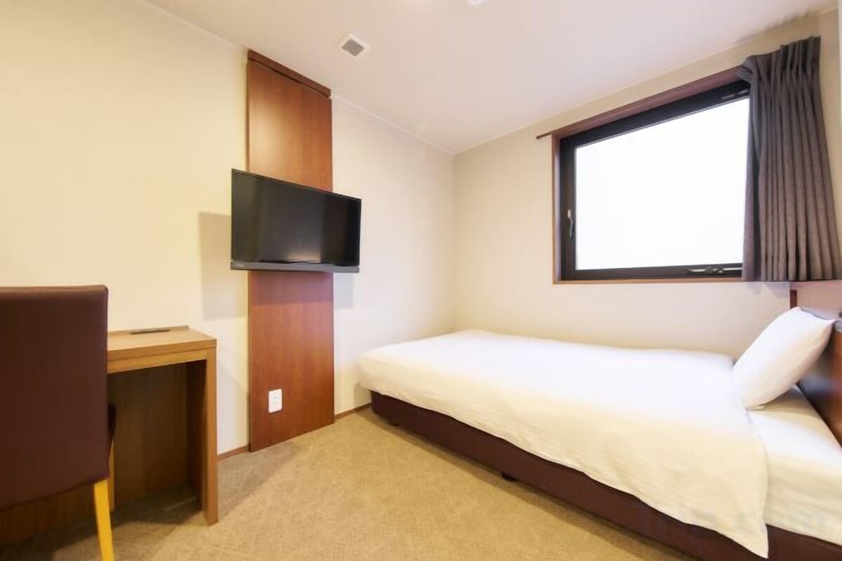 A small, single-bed room with a television mounted on the wall, a desk, and a window with curtains.