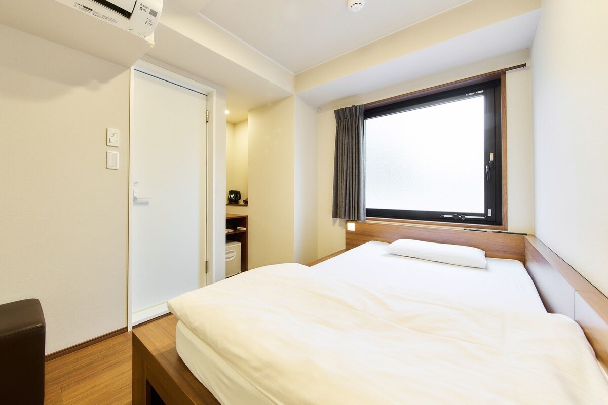 A hotel room with a bed, a desk, a chair, a window with curtains, and a door.