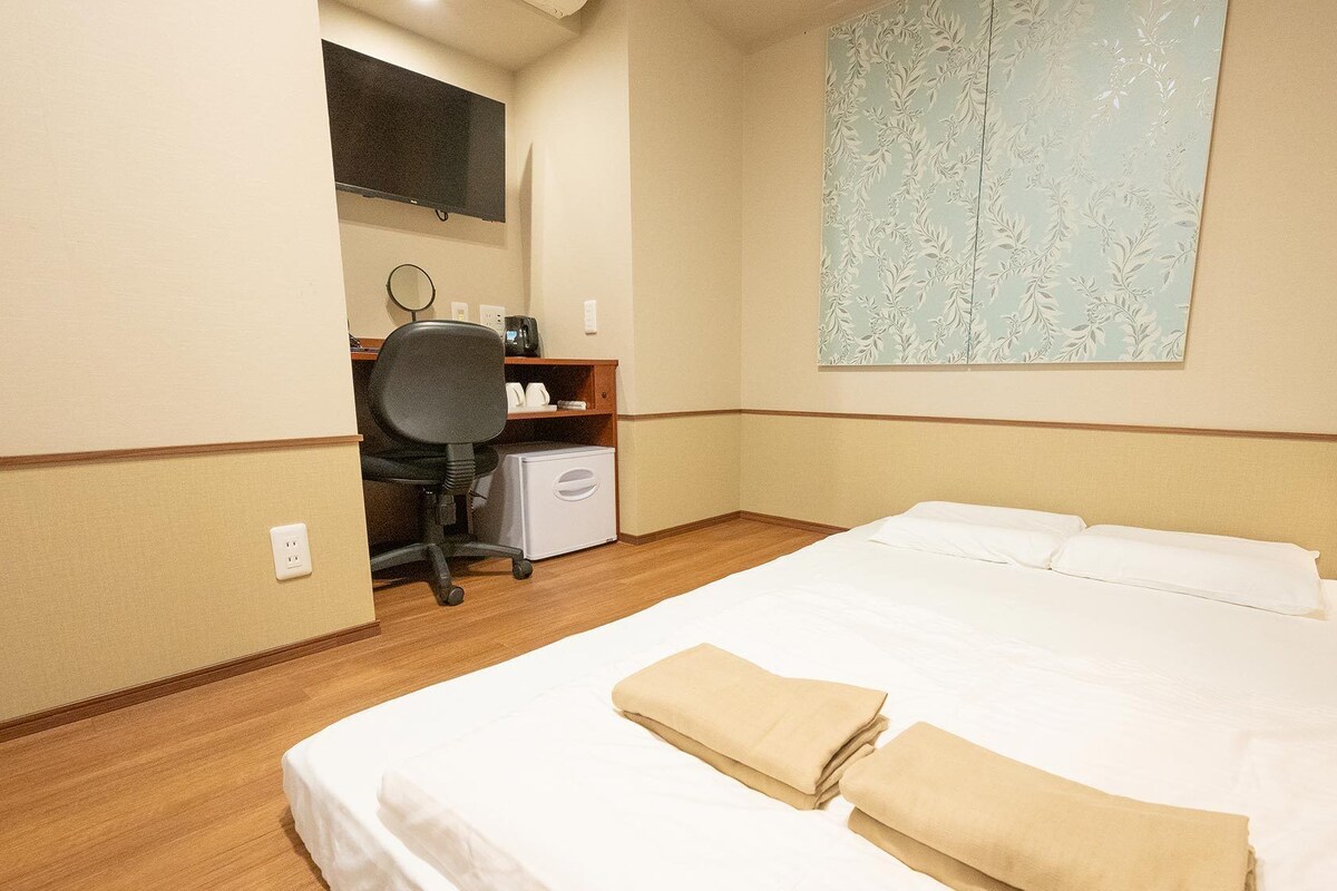 A hotel room with a bed, a desk, a chair, a small refrigerator, a mirror, and a wall-mounted TV.