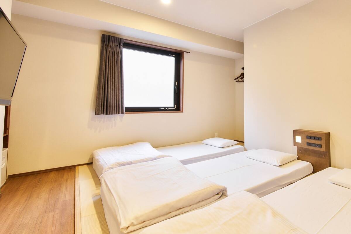 Habitación de hotel con tres camas individuales, una ventana con cortinas, un cabecero de madera y una mesita de noche pequeña.