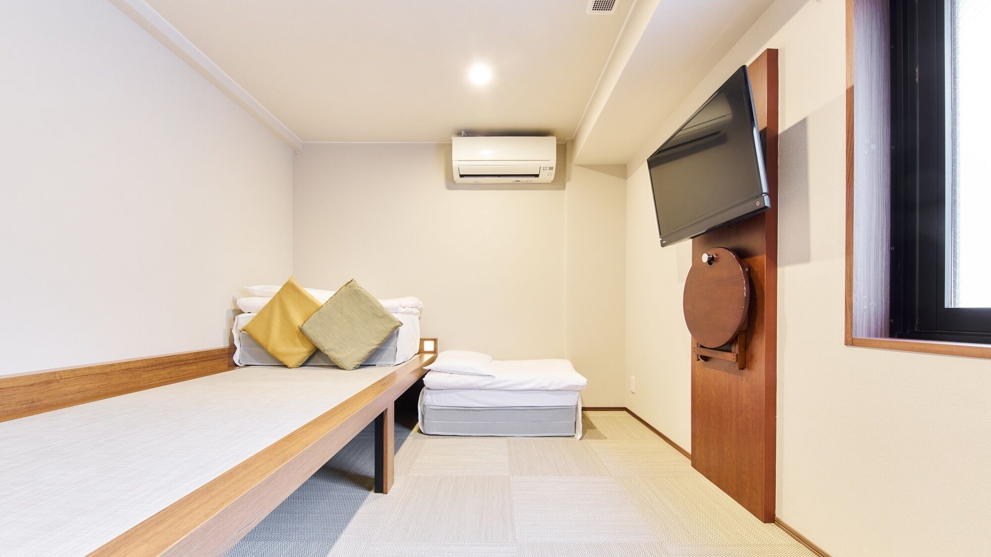 A small, single-room hotel room with a bed, a TV mounted on the wall, and an air conditioning unit.