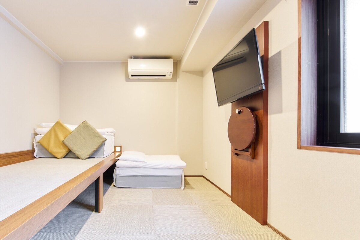 A small, single-room hotel room with a bed, a TV mounted on the wall, and an air conditioning unit.