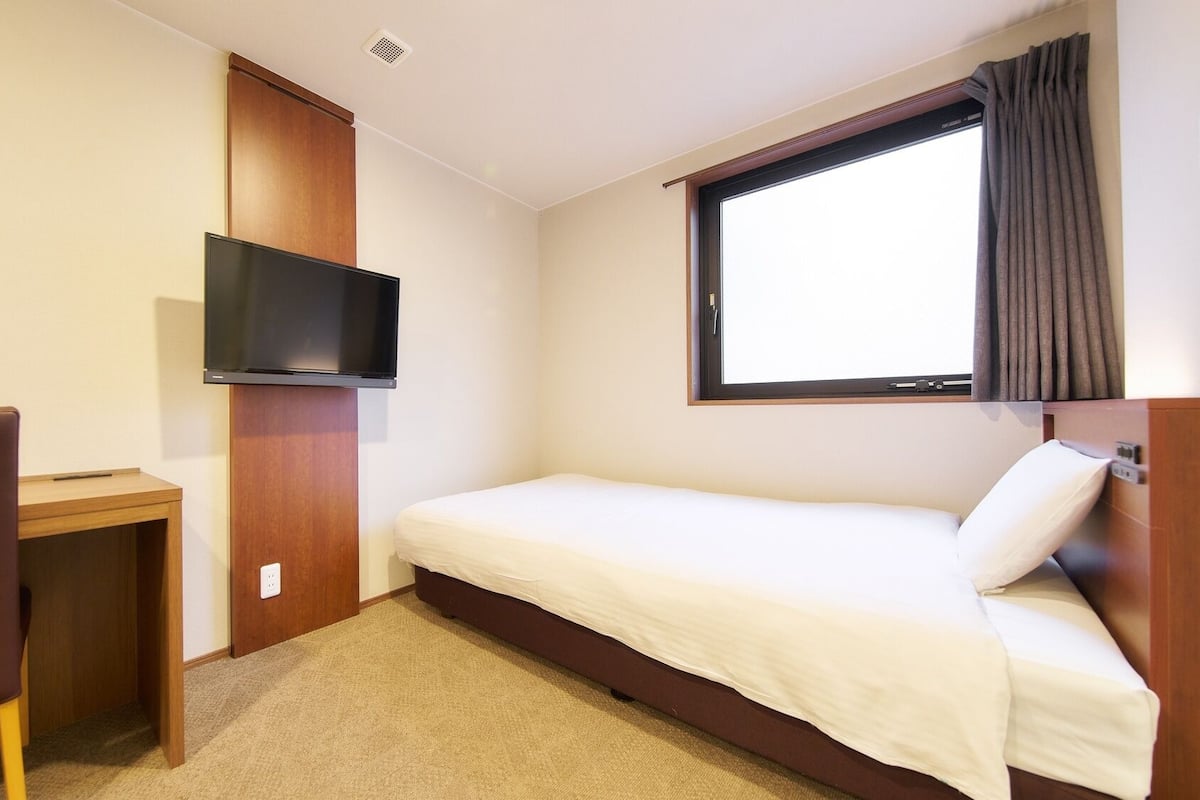 Una habitación pequeña con una cama individual, televisión montada en la pared, escritorio y ventana con cortinas.