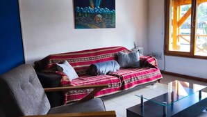 Sala de estar com teto de madeira, sofá com almofadas estampadas, mesa de centro de vidro e pintura de uma paisagem nevada.