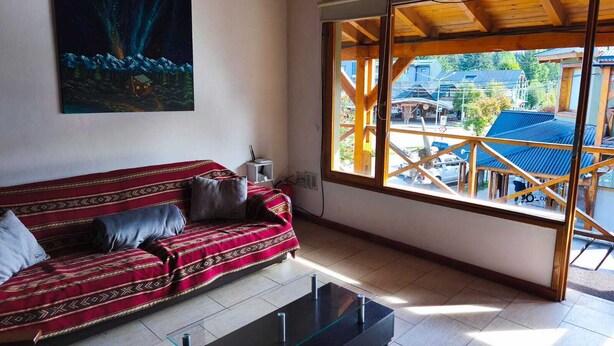 Uma sala de estar com um sofá vermelho estampado, uma mesa de centro de vidro e uma grande janela com vista para prédios e árvores.