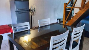 Uma sala de jantar com mesa de madeira e cadeiras brancas, geladeira e ventilador.