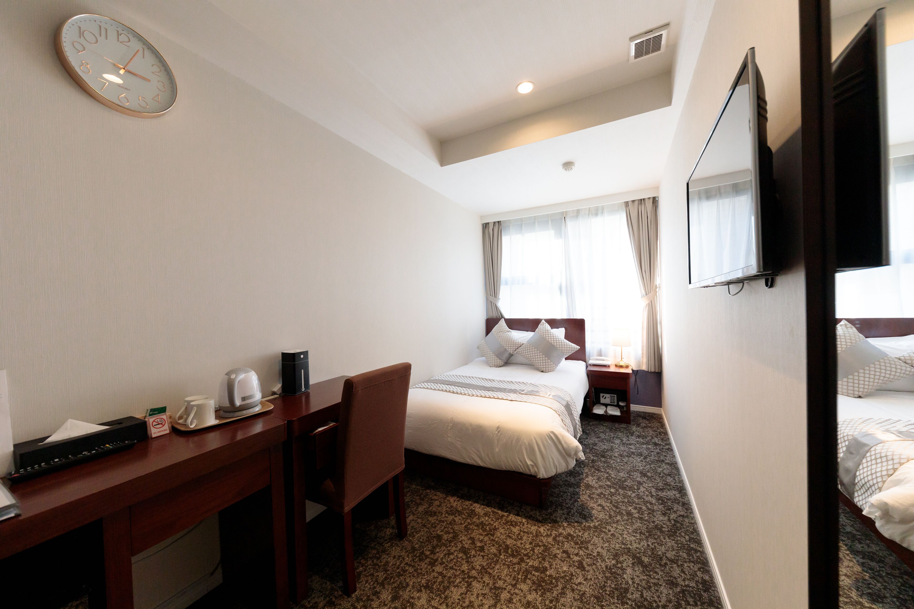 A hotel room with a bed, a desk with a chair, a clock, a television, and a window with curtains.