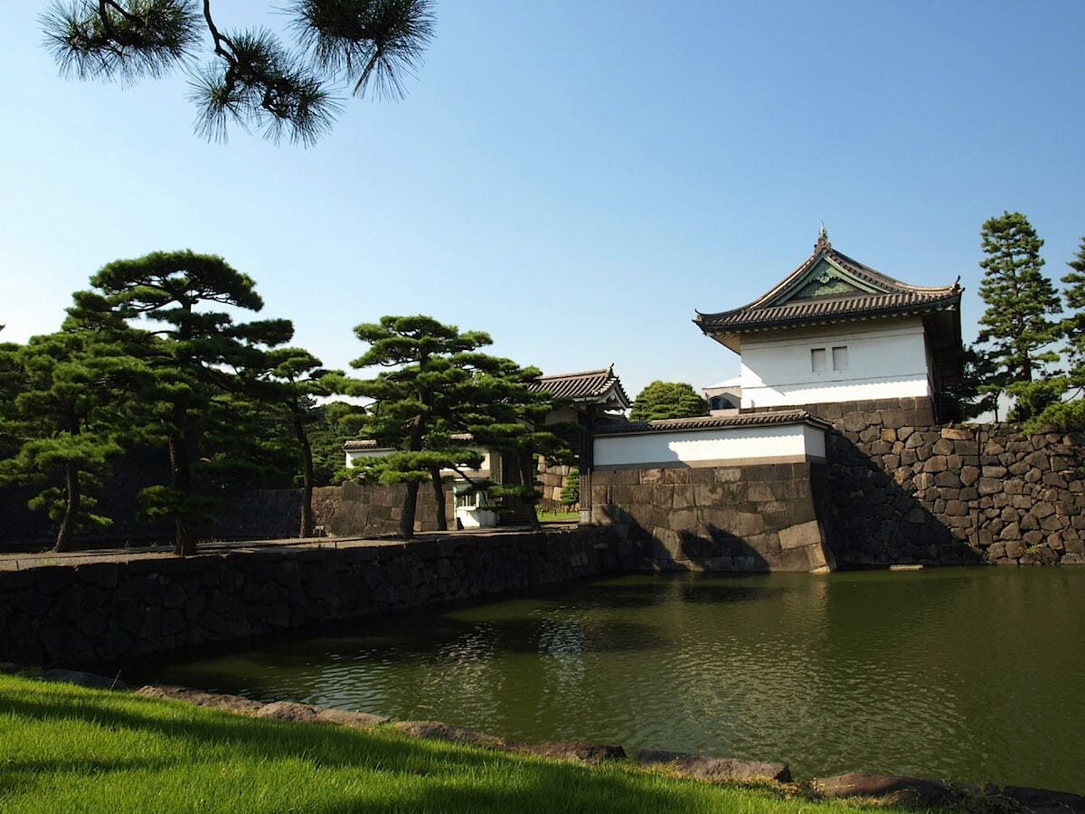 Un bâtiment traditionnel japonais, doté d’un toit recouvert de tuiles, entouré d’un mur de pierre et d’un petit étang.