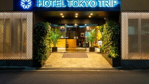 The entrance of Hotel Tokyo Trip with a modern design, featuring a prominent illuminated sign, a glass door, and potted plants. - HOTEL Tokyo Trip Ueno Nishi Nippori (Tokyo)