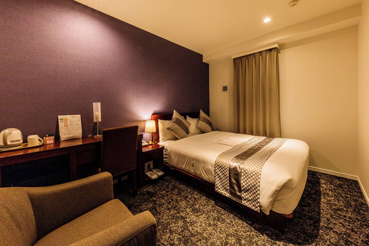 A hotel room with a bed, a desk with a lamp, a chair, and a small table with a phone and a menu.