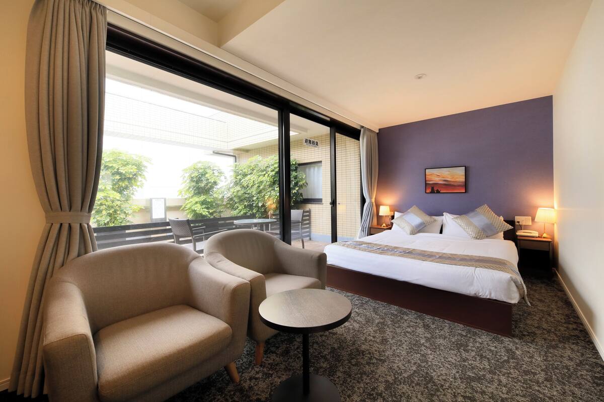 A hotel room with a bed, two armchairs, a small table, and a view of the outdoors.