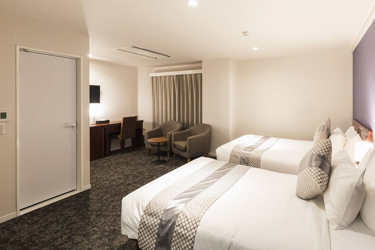 A hotel room with two beds, a desk, a chair, and a window with curtains.